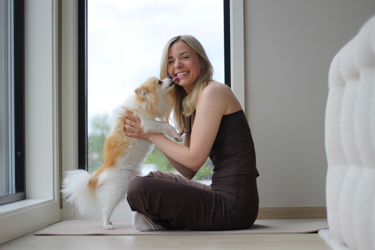 Malin Kristine Moen sitter med korslagte ben på en treningsmatte ved et stort vindu og smiler mens den lille hunden hennes slikker henne på kinnet.